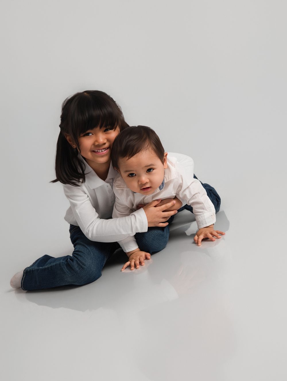 Young girl smiling and hugging a baby sibling on a white studio floor, both wearing white tops and jeans.