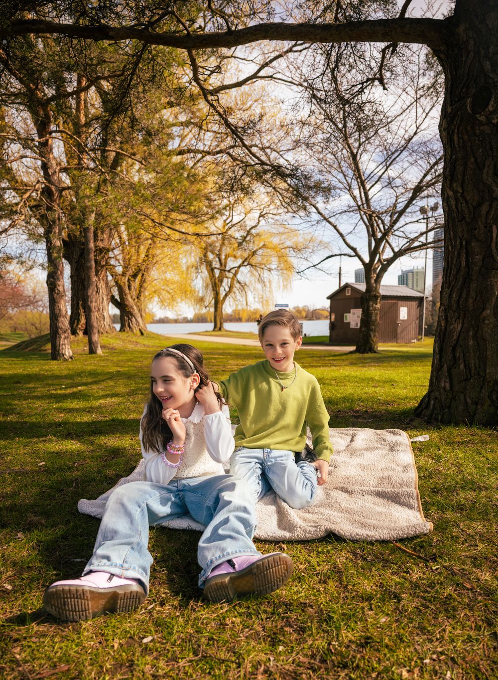 Two children sit on a blanket under trees in a sunny park near a lake, one in white and one in green.
