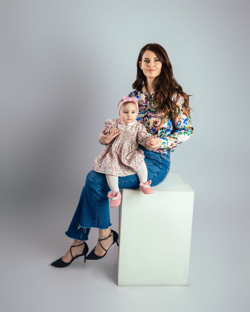 Mother in floral blouse and jeans sitting on white cube with baby girl in pink outfit.