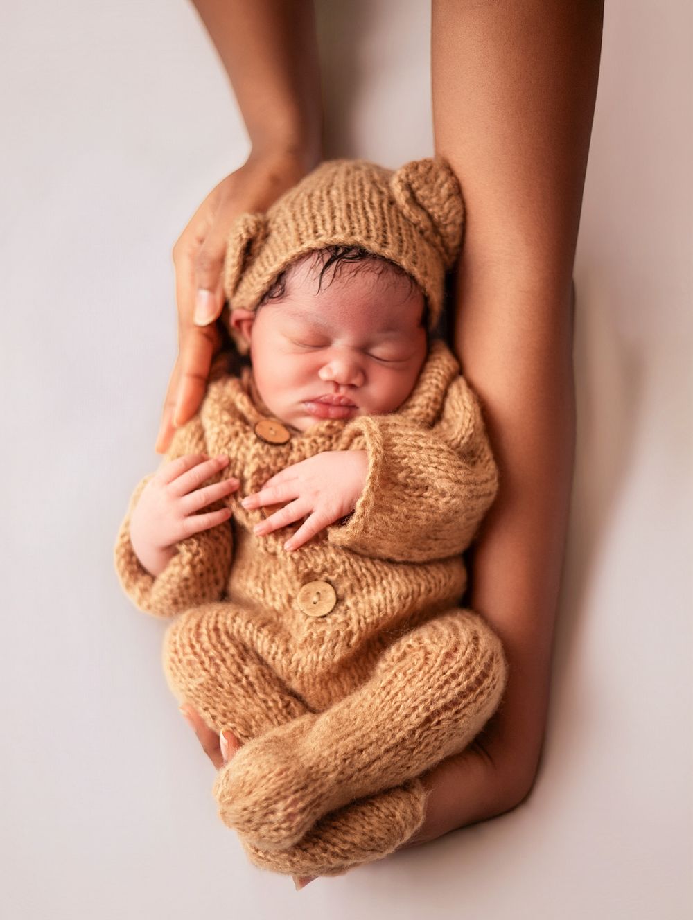 Newborn baby sleeping peacefully in a tan knit bear outfit, cradled gently in an adult's hands.