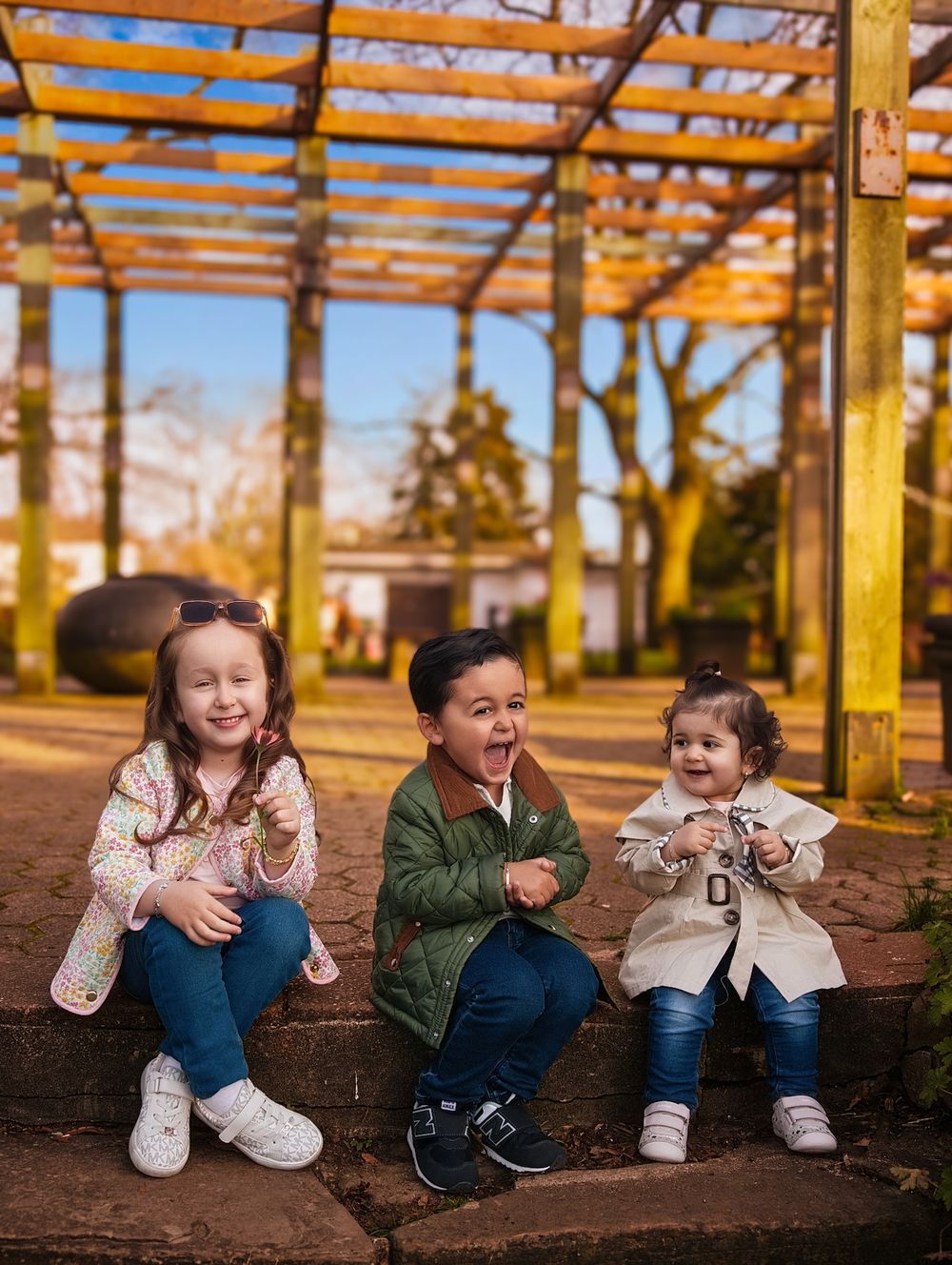 Three happy children sitting outdoors under a rustic pergola in a sunny park, wearing colorful jackets and jeans.