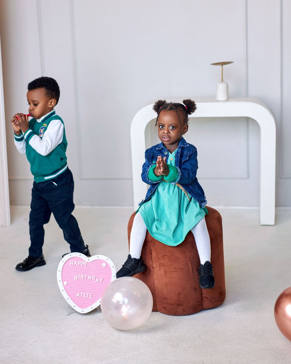 Two young children in stylish outfits celebrate a birthday with balloons and a heart sign.
