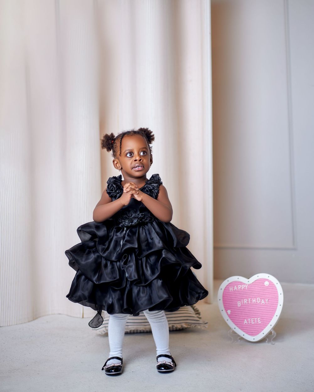 Little girl in a black ruffled dress stands beside a Happy Birthday heart sign.