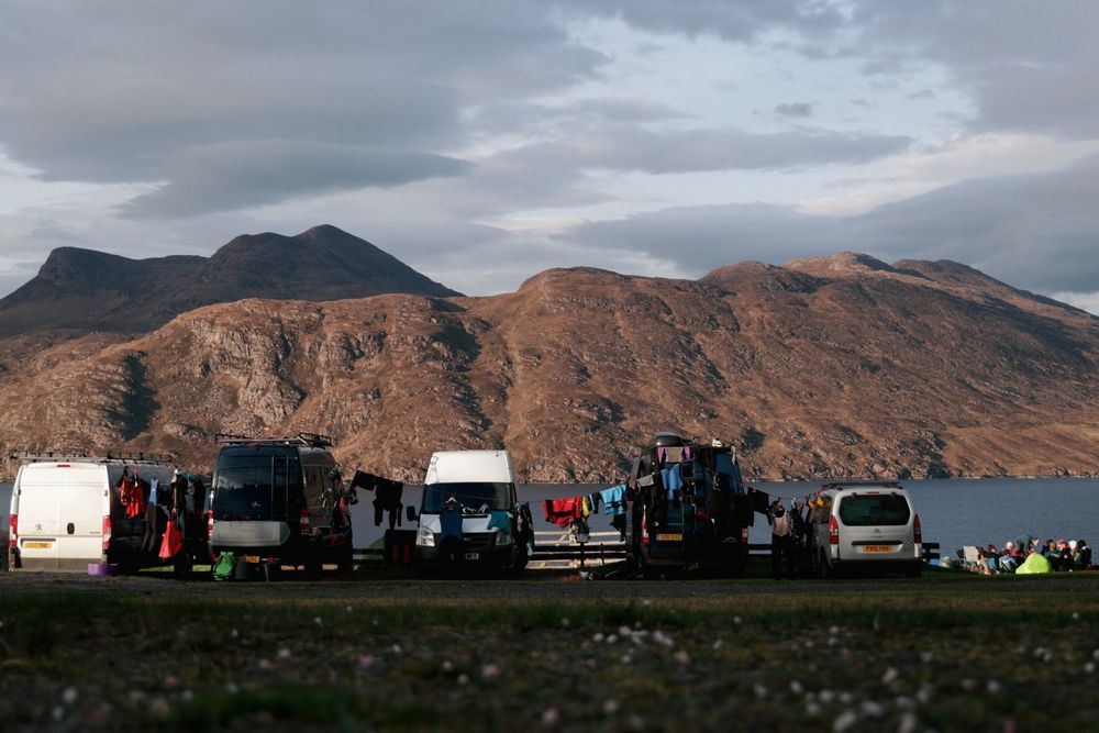 A panoramic view of campervans with wetsuits hanging parked against a backdrop of rugged mountain peaks and cloudy skies.