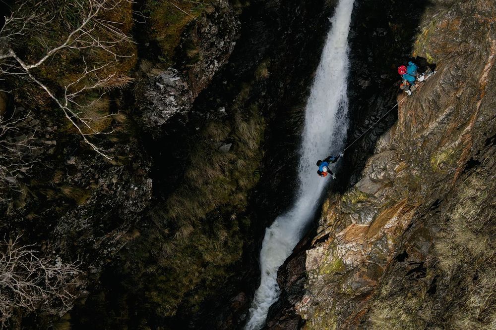 A canyoneer abseils down a tall cascading waterfall with in a scenic mountain landscape.