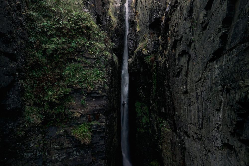 A cascading waterfall flows down a dark rocky cliff face surrounded by lush green vegetation in a forest setting.