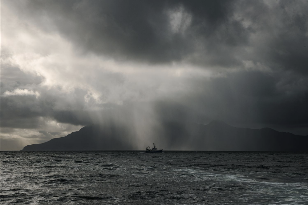 A small boat sails beneath dramatic storm clouds and rain shafts over dark ocean waters.