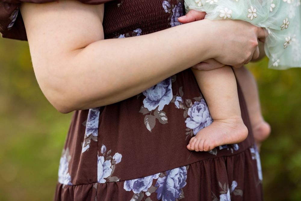 Close up view of bare baby feet being held against a brown floral dress in an outdoor setting.