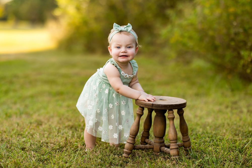 A small child in a floral dress plays with a wooden stool outdoors in a sunlit garden setting.