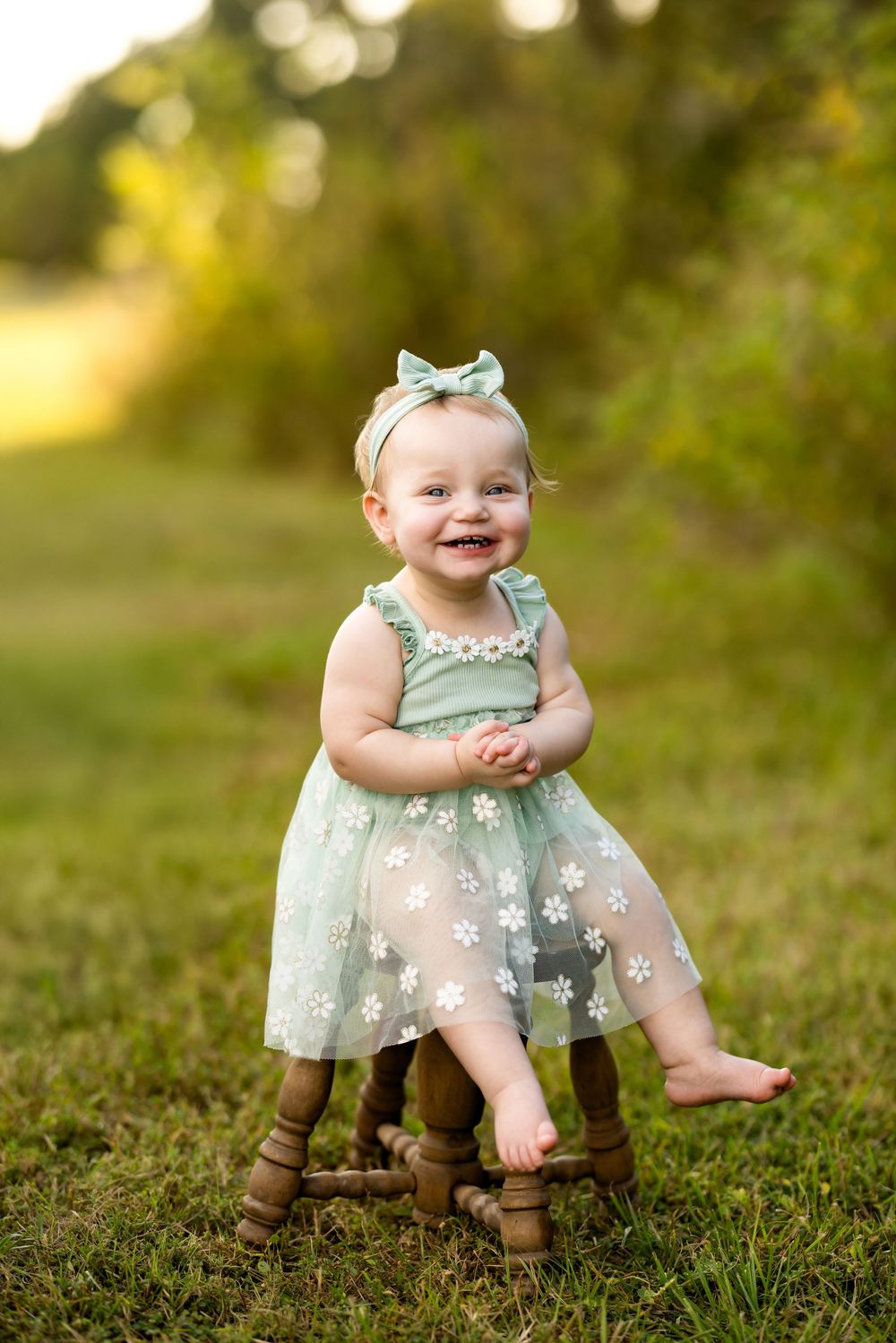 A small child in a mint green tulle dress sits on a wooden stool in a grassy outdoor setting.
