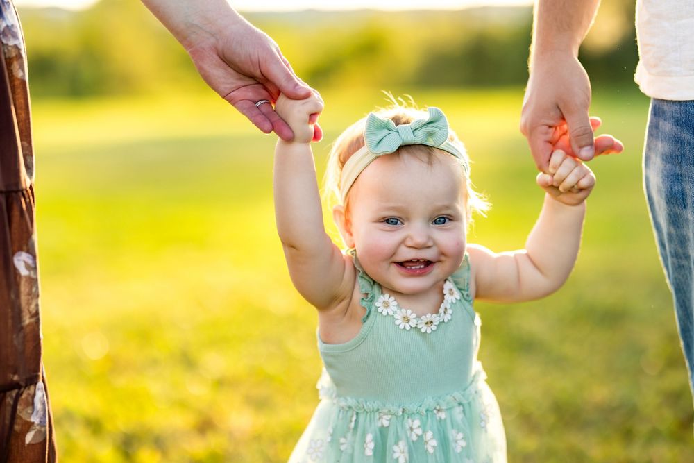 Baby in mint green dress smiles while holding hands with adults in sunny outdoor setting.