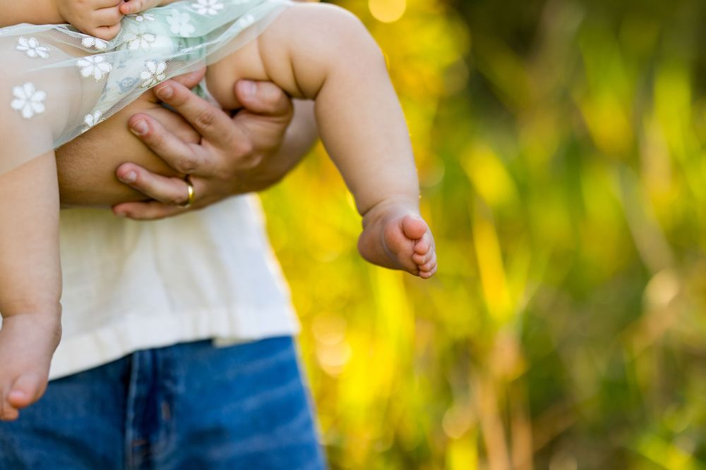 Small bare feet and legs of a toddler wearing jeans against a soft yellow and green blurred nature background.