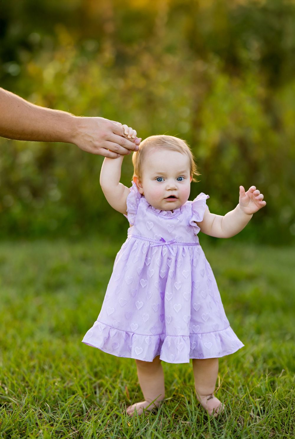 A small child in a lavender ruffled dress stands in grass while a hand reaches to steady them from above.