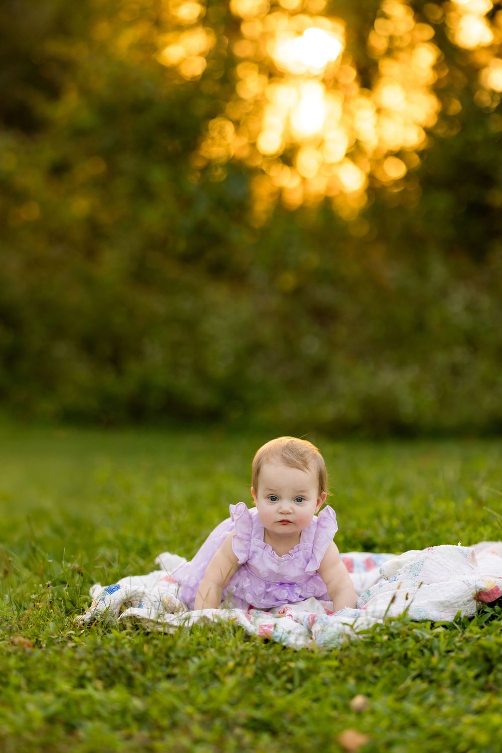 Baby in pink dress lies on white blanket in grassy field during golden sunset.