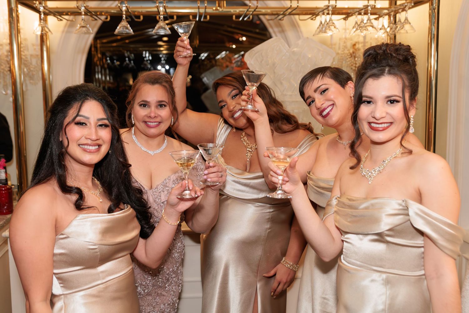 Five women in gold satin dresses celebrating and holding champagne glasses at formal event.