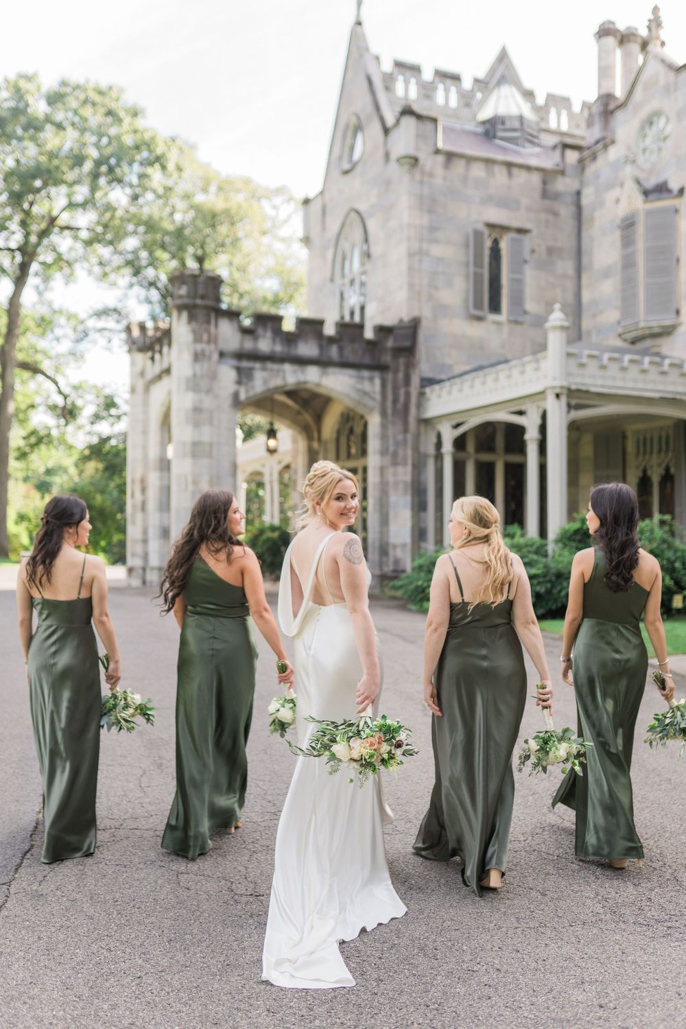 Bridesmaids in dark green dresses walking with bride in white gown at gothic stone castle venue.
