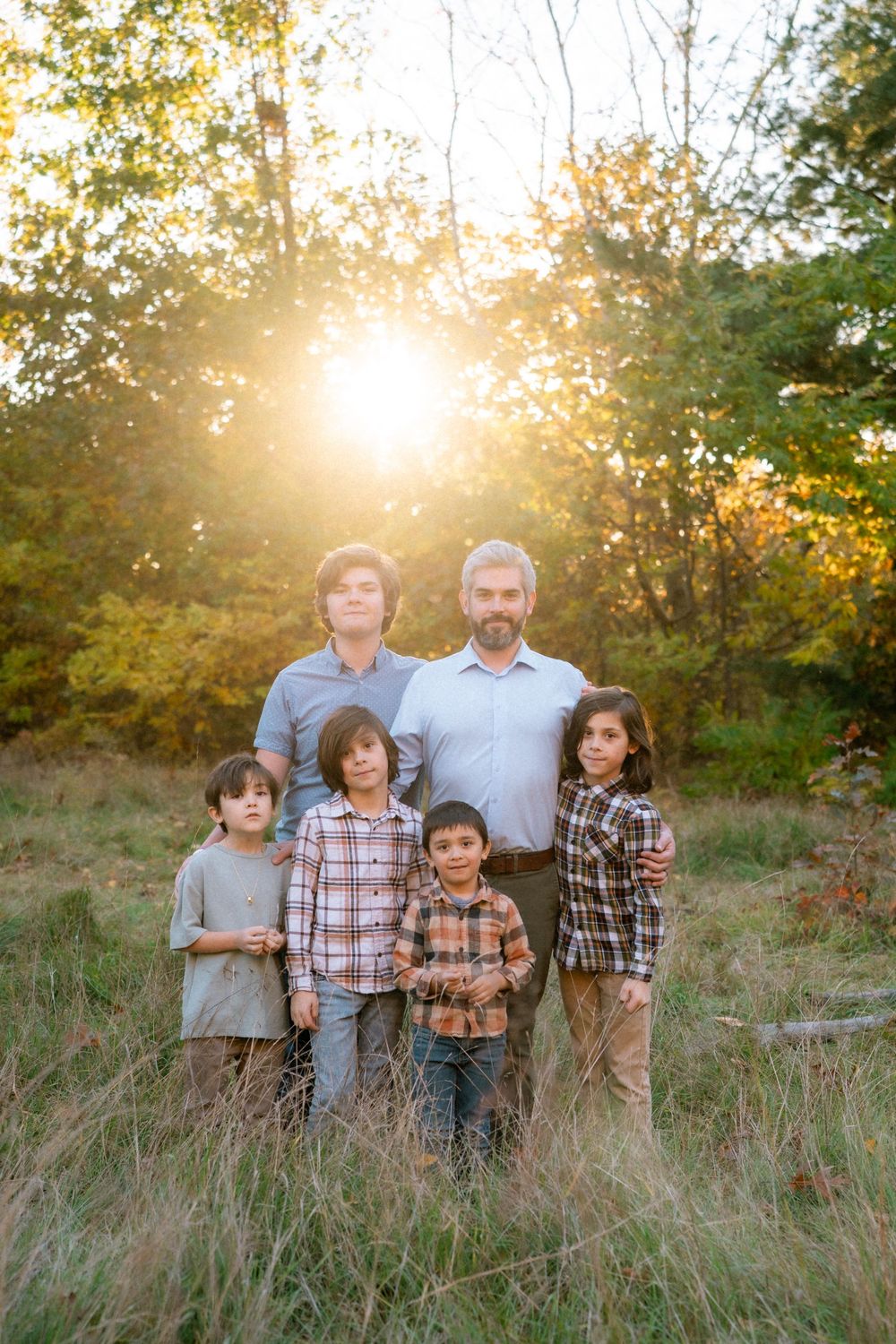 A family poses together in a sunlit field during golden hour while wearing coordinating plaid and casual attire.