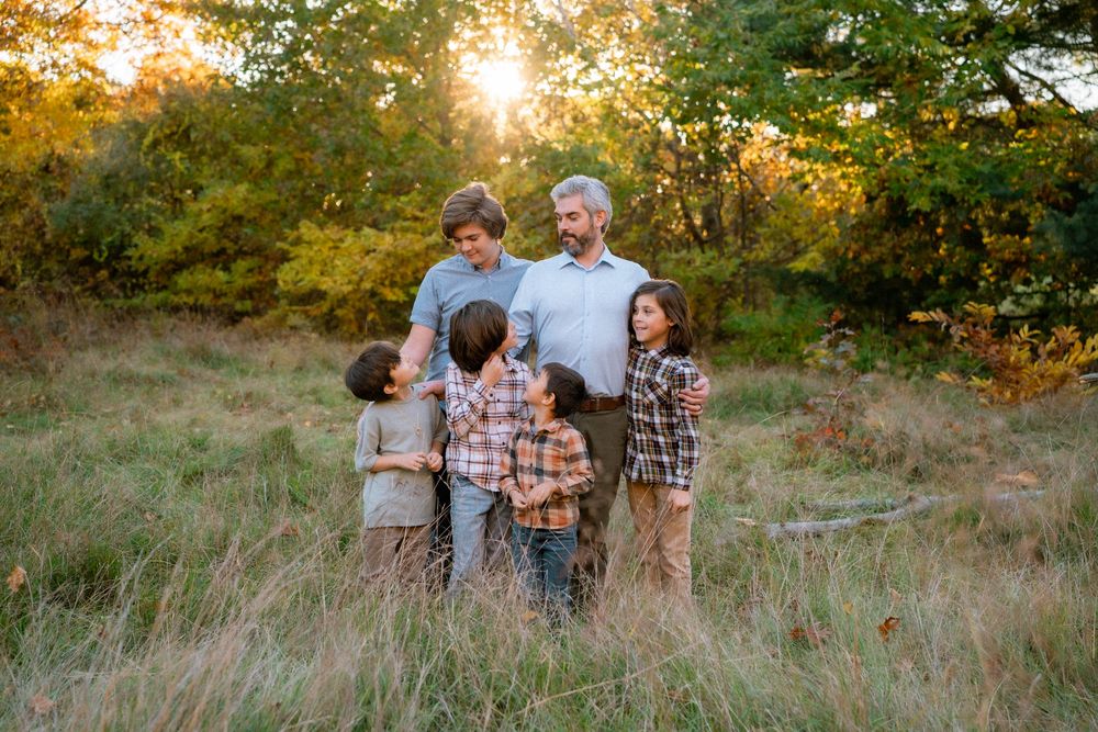 A family of five stands together in a grassy field at sunset with golden sunlight filtering through trees behind them.