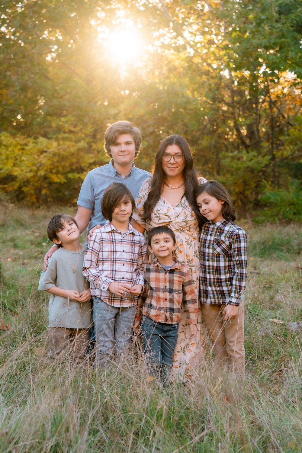 A family poses together in a sun-drenched field with golden light filtering through trees during sunset.