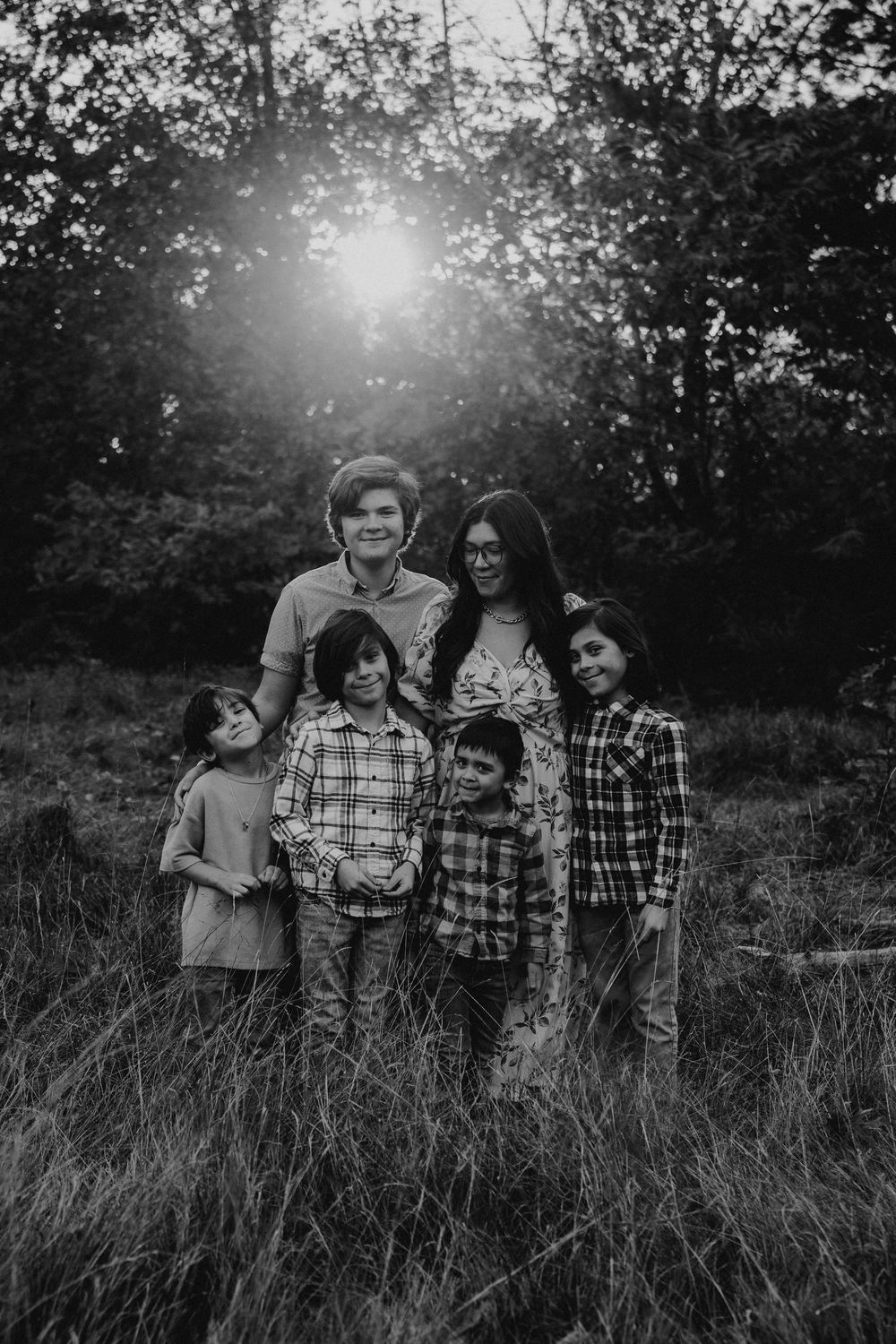 Family members embrace while standing in a grassy field with sunlight filtering through trees behind them.