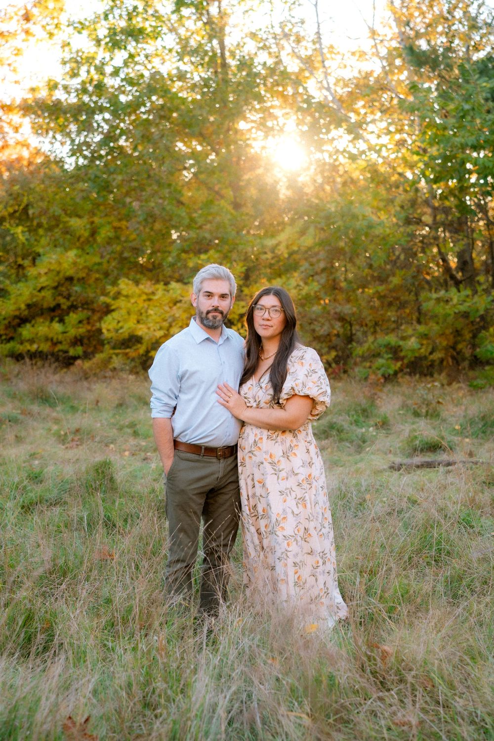 A couple embraces in a golden sunlit field with tall grass and autumn foliage during sunset.