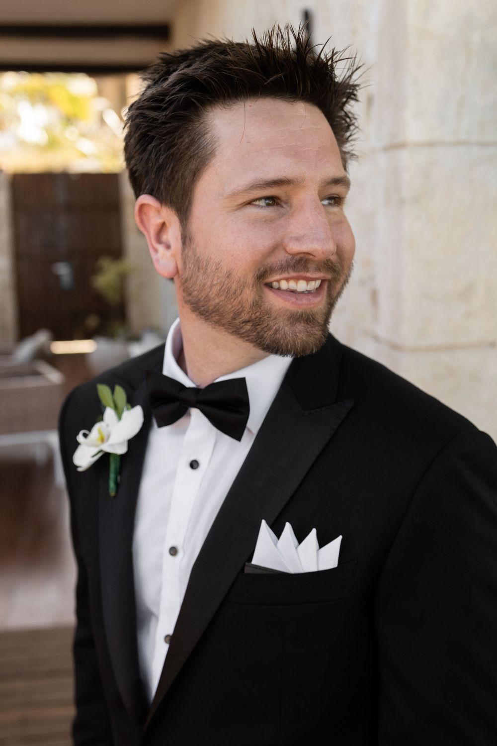 Groom wearing classic black tuxedo with white bow tie and boutonniere smiling in an elegant indoor setting.