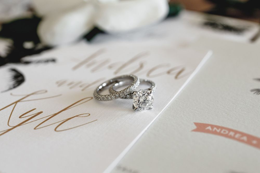 Wedding rings with diamond accents rest on a white invitation card with elegant script lettering.