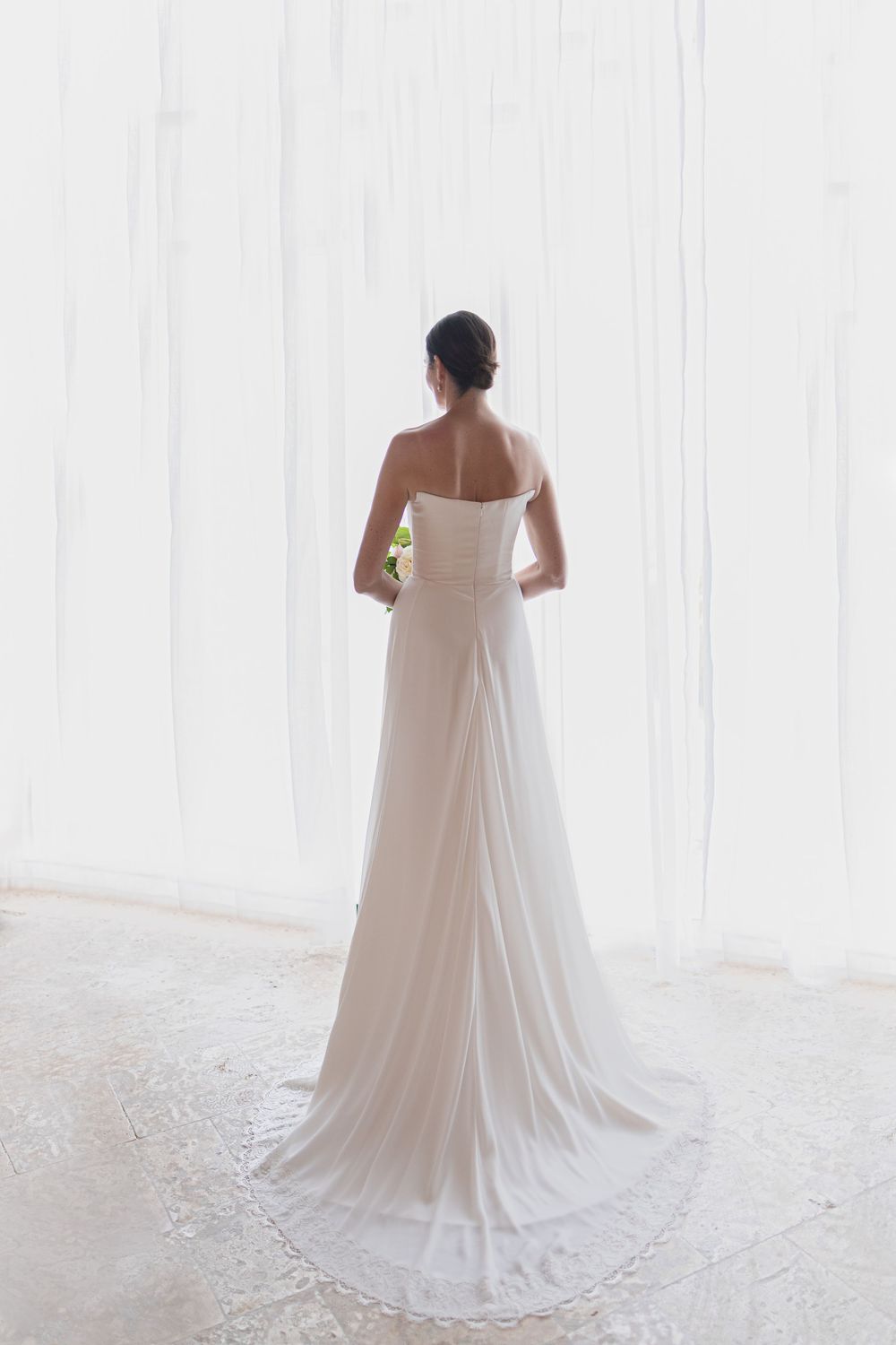 Elegant white wedding dress with flowing train photographed from behind on white background.