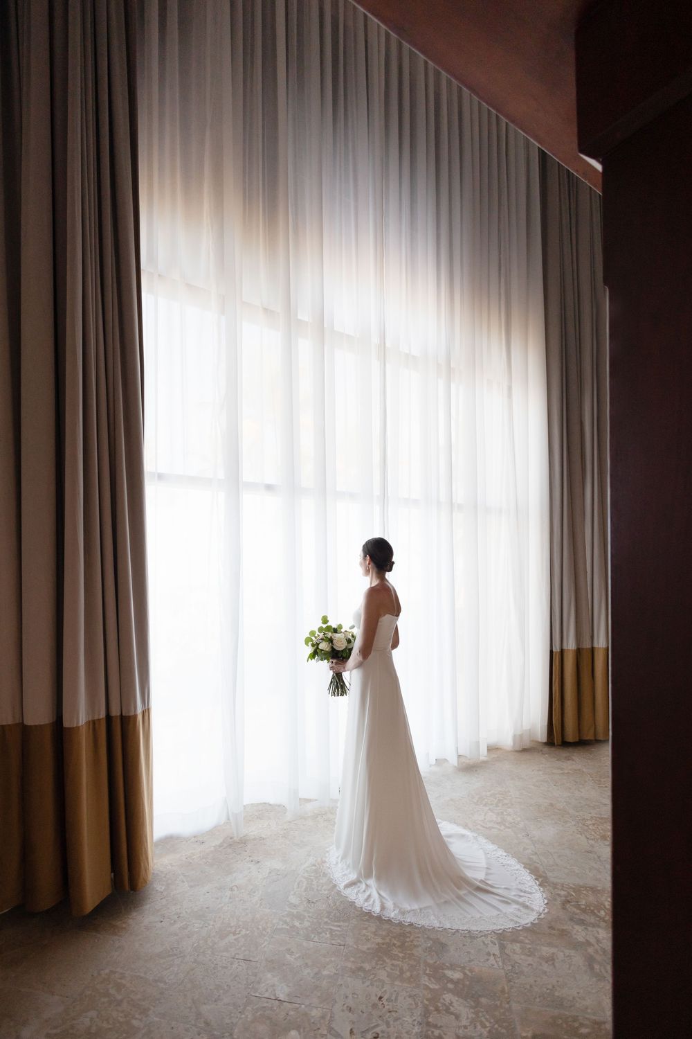 Dramatic silhouette of a bride against sheer curtains in a minimalist indoor setting.