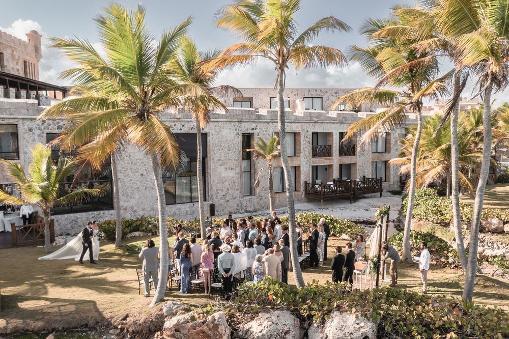 Wedding guests seated in rows of white chairs on a palm-lined beachfront during an outdoor ceremony.