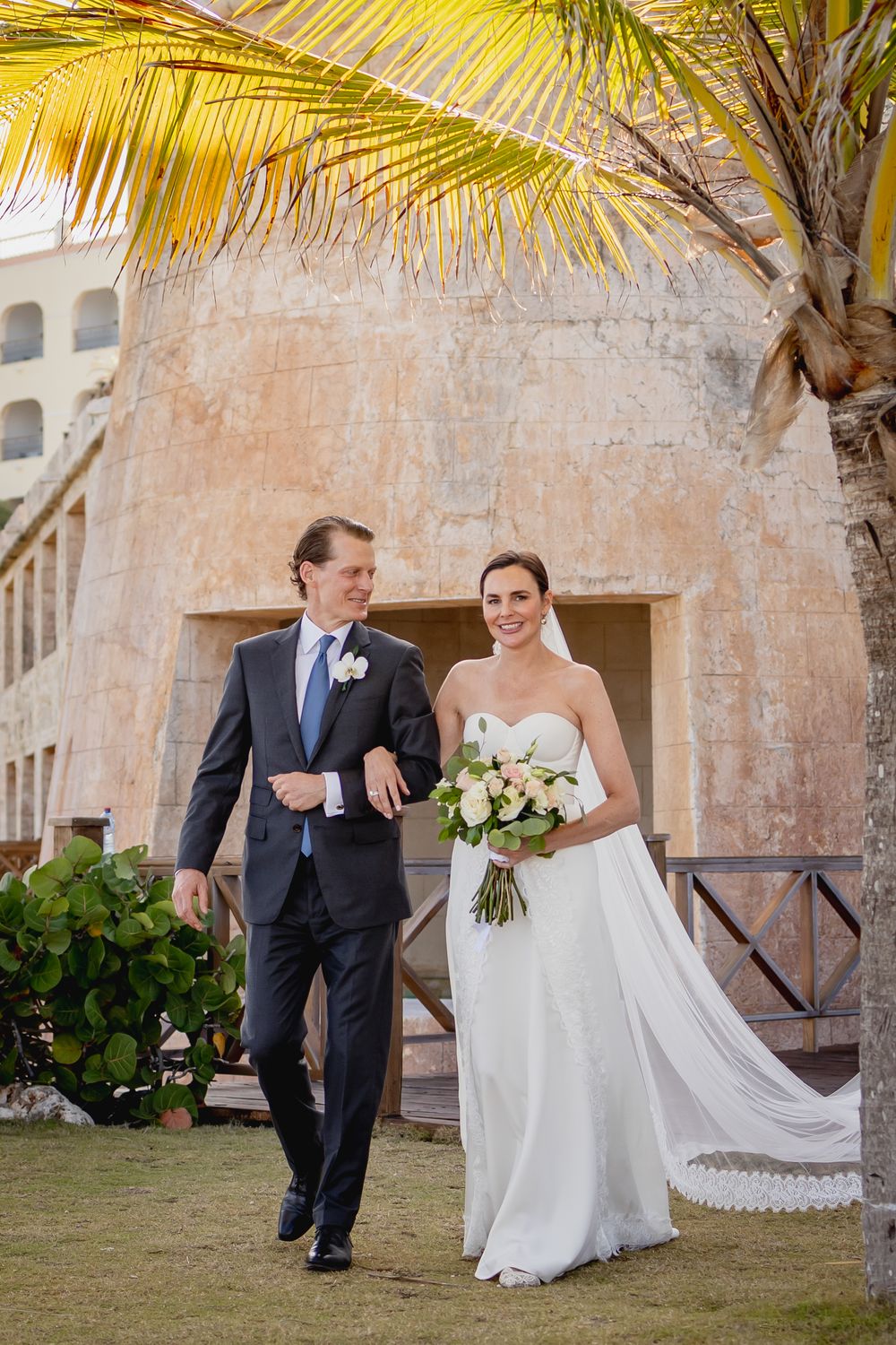 A wedding couple poses beneath swaying palm trees and stone architecture at a tropical venue.