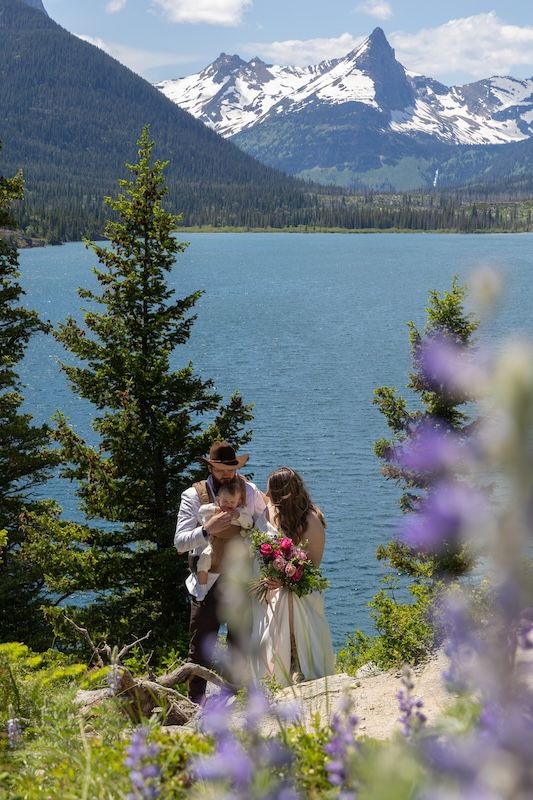 Couple exchanging vows at lakeside elopement with snow-capped mountains, wildflowers, and turquoise water in background.