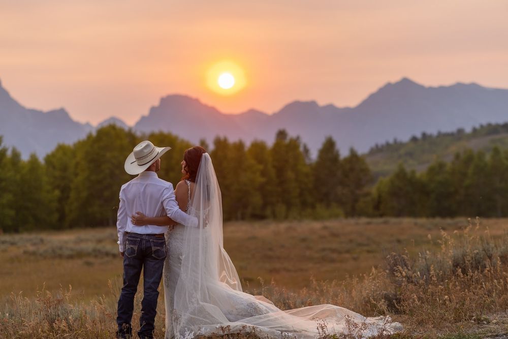 A couple in western wedding attire watches the sunset against a mountain backdrop in a rustic field setting.