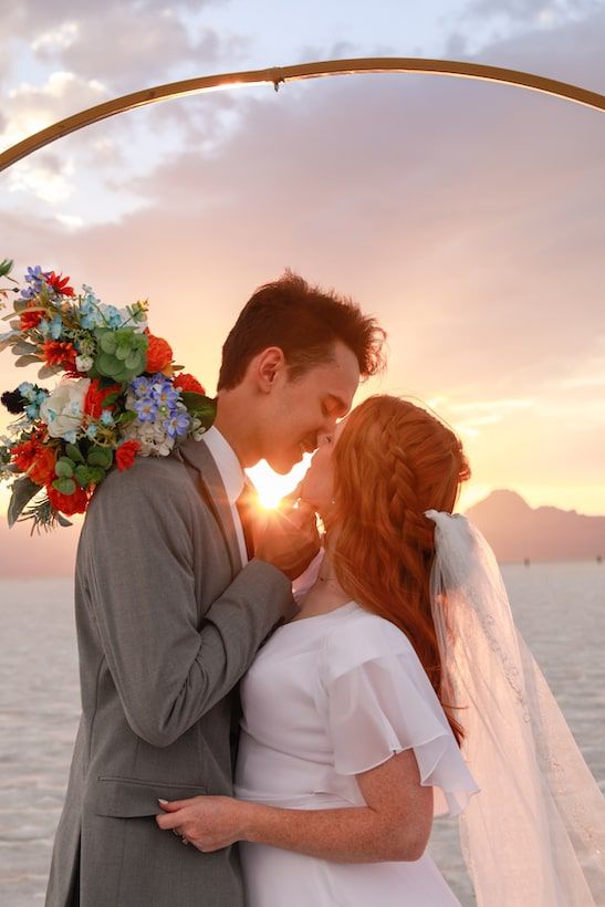 A romantic couple shares tender moments at sunset by the ocean with floral decor and flowing veil.