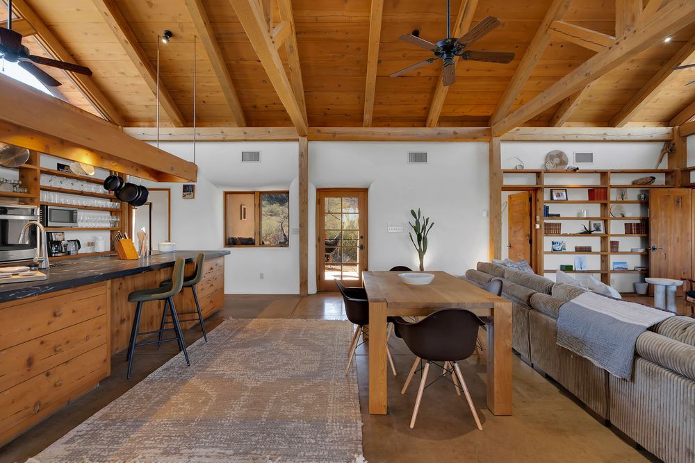 Modern open-concept living space with exposed wooden beams, gray sectional sofa, and natural light flooding through windows.
