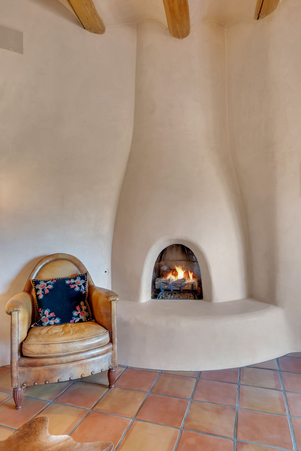 Adobe-style curved walls and wooden beams create a cozy southwestern interior with terracotta tile floors and built-in seating.