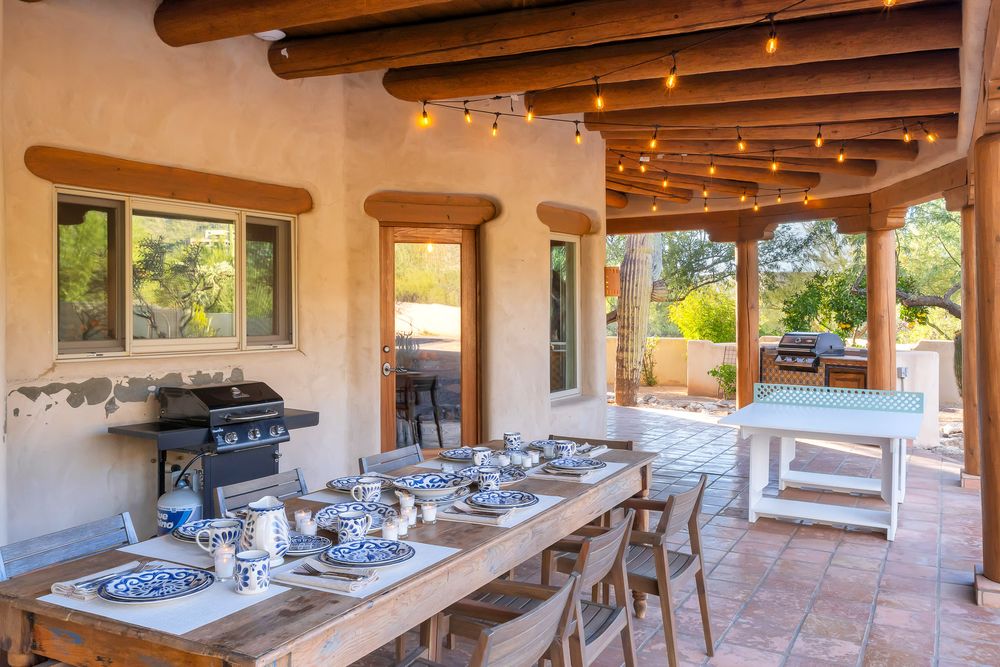 Rustic covered patio with exposed wooden beams and outdoor dining furniture set against adobe-style architecture.