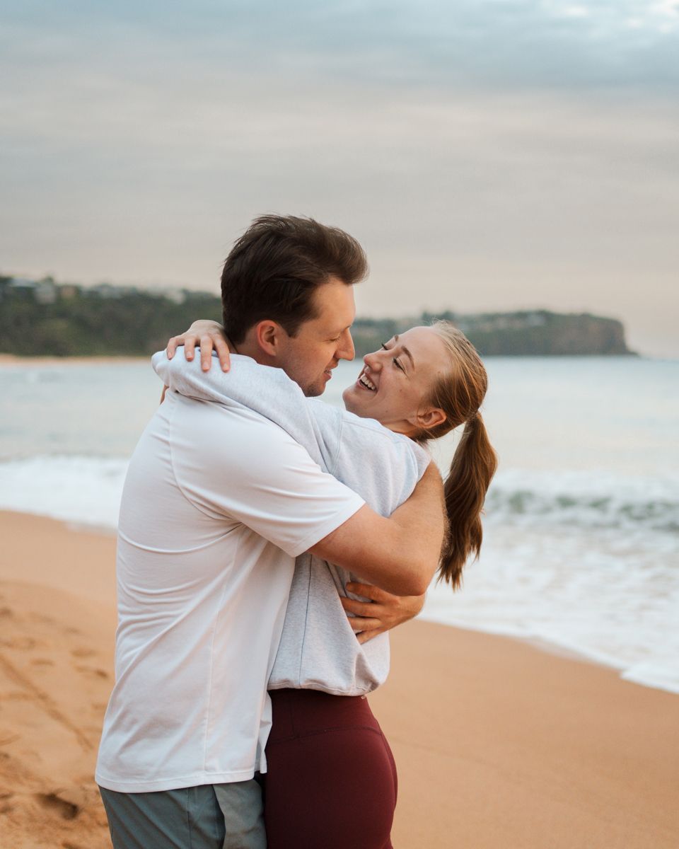 couple hugging and looking at each other Newport beach with ocean waves and cliffs in the background at sunset during engagement photoshoot