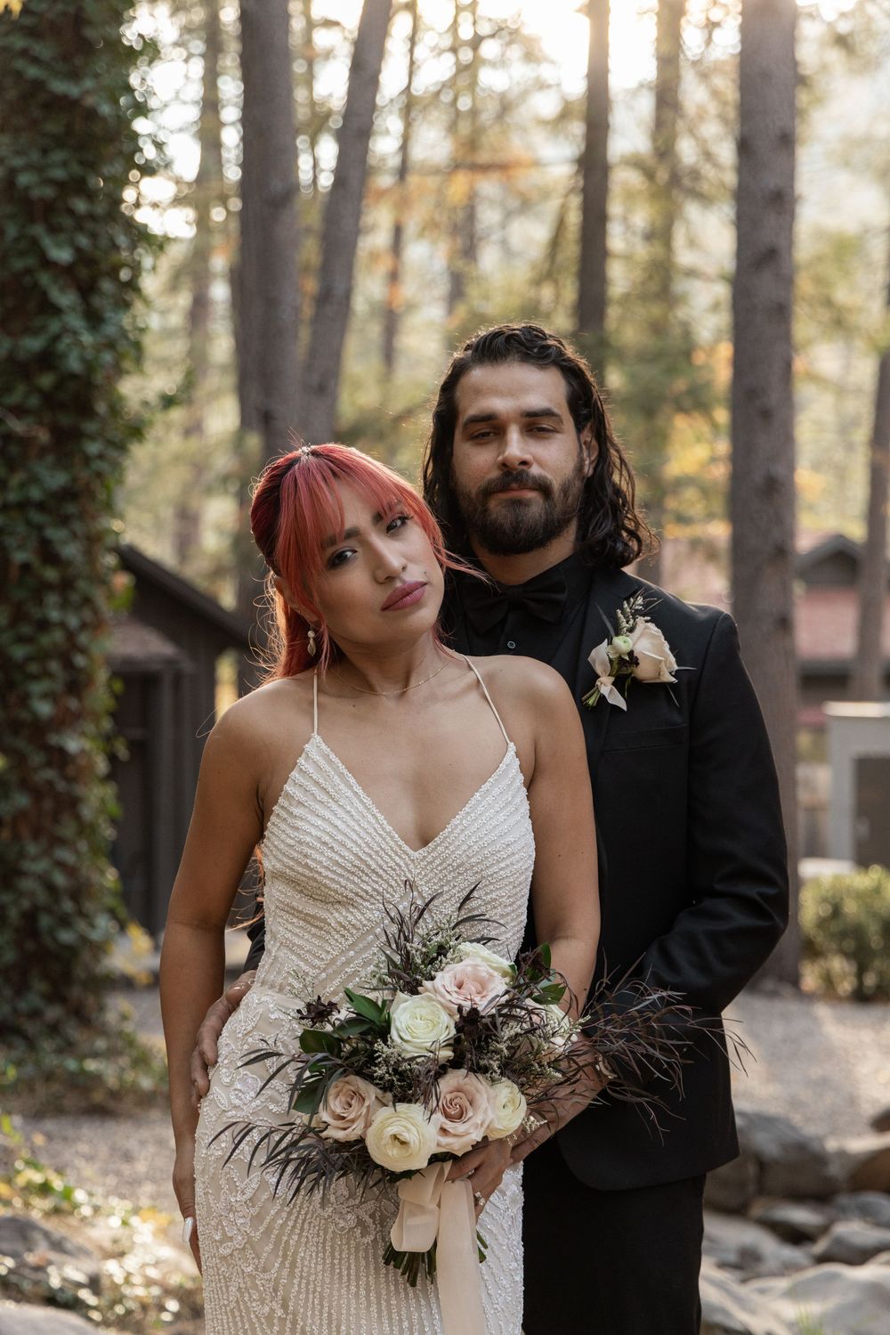 A couple poses together in a wooded setting wearing wedding attire with the bride holding a romantic floral bouquet.