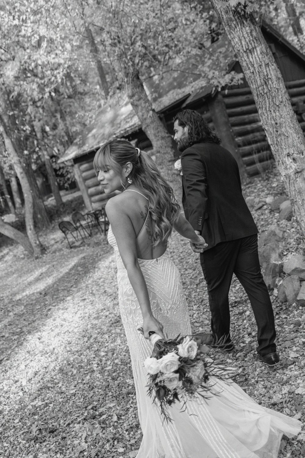 A couple in wedding attire poses for romantic photos in a wooded outdoor setting with fallen autumn leaves.