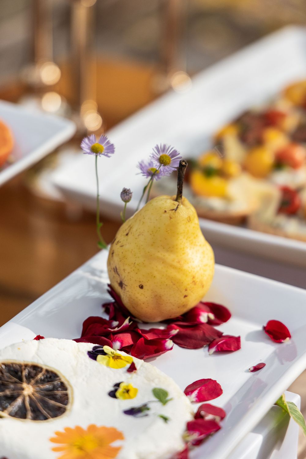 Elegant plated dessert with yellow sorbet ball garnished with purple flowers and red rose petals on white dish.