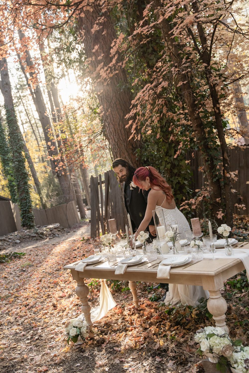 A rustic outdoor table setting in an autumn forest with white linens and place settings surrounded by fallen leaves.