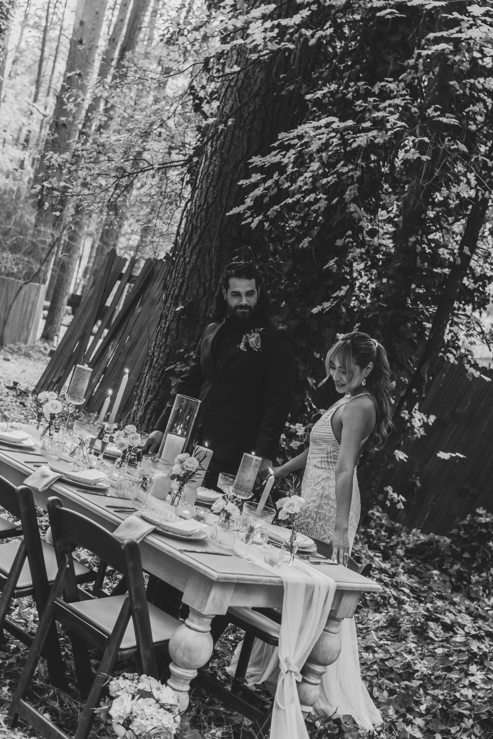 Black and white photograph showing an elegant outdoor dinner party setup in a wooded forest setting.