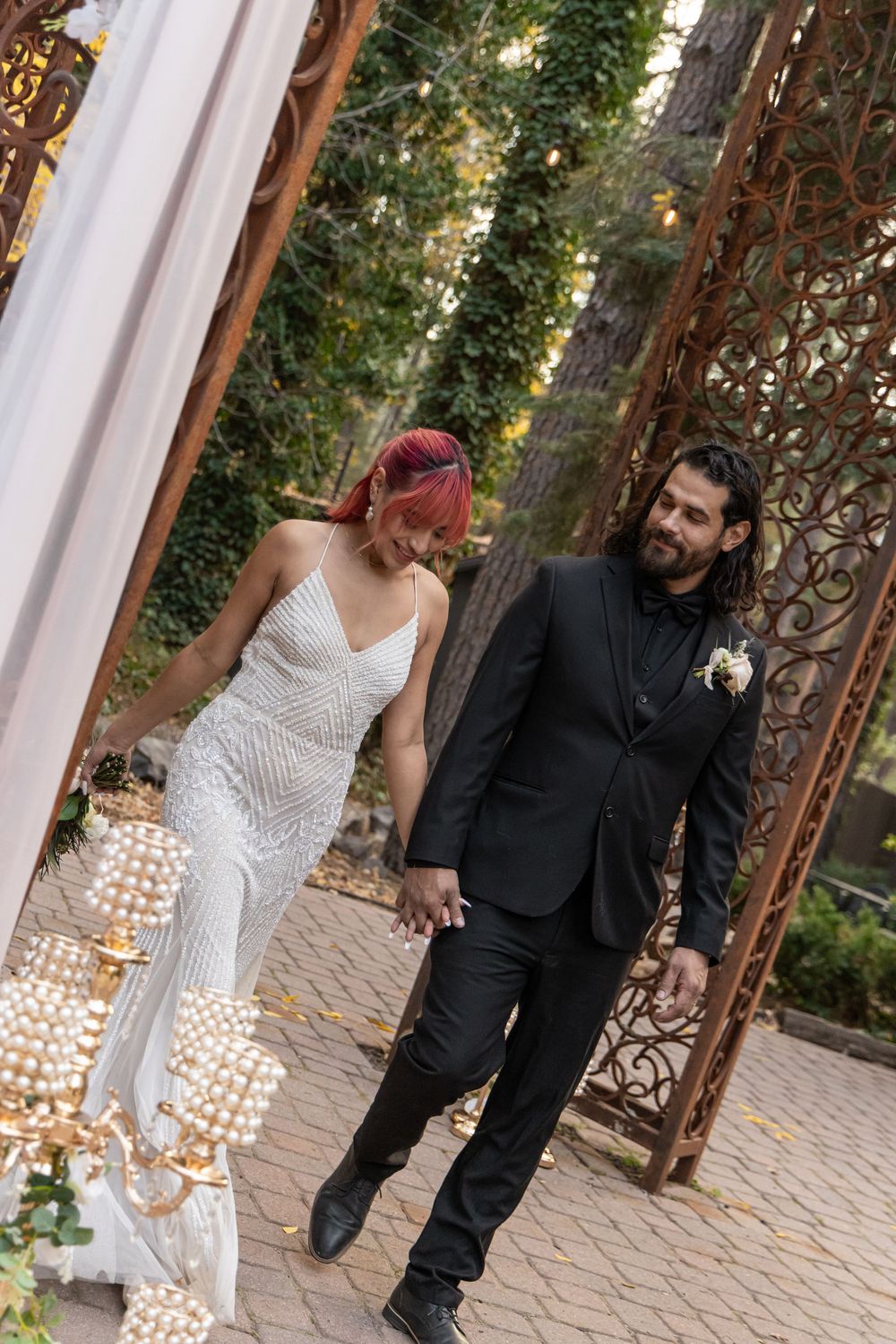 A couple walks together under a white archway decorated with glowing lights in a garden setting.