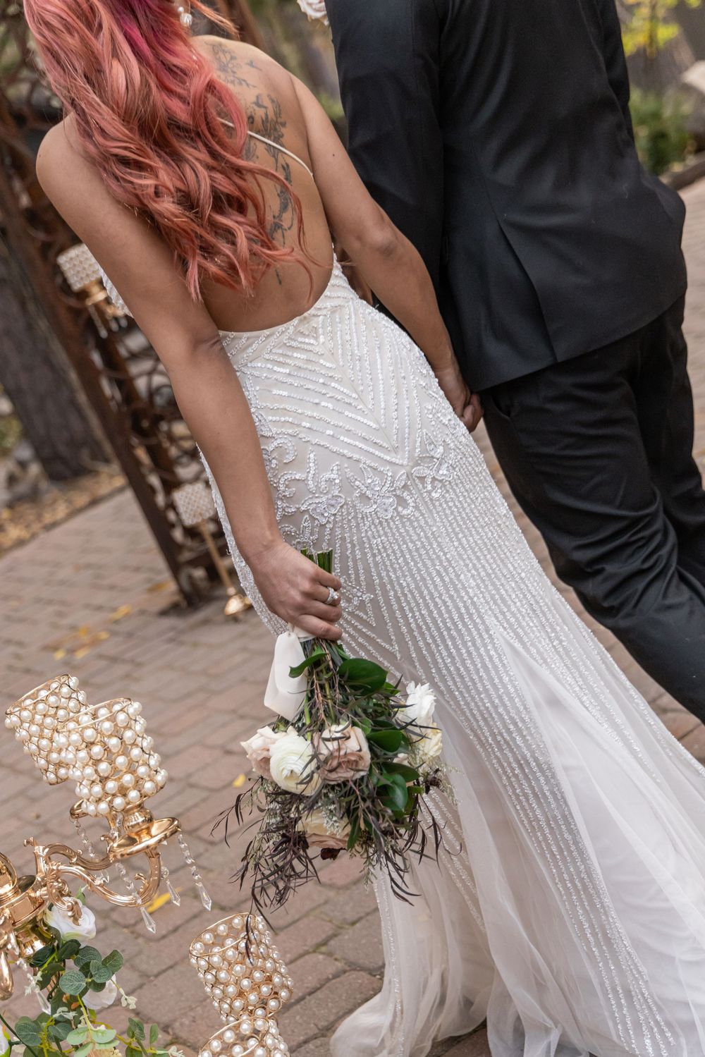 A couple in wedding attire walking together, with the bride wearing a beaded white dress and holding a small bouquet.