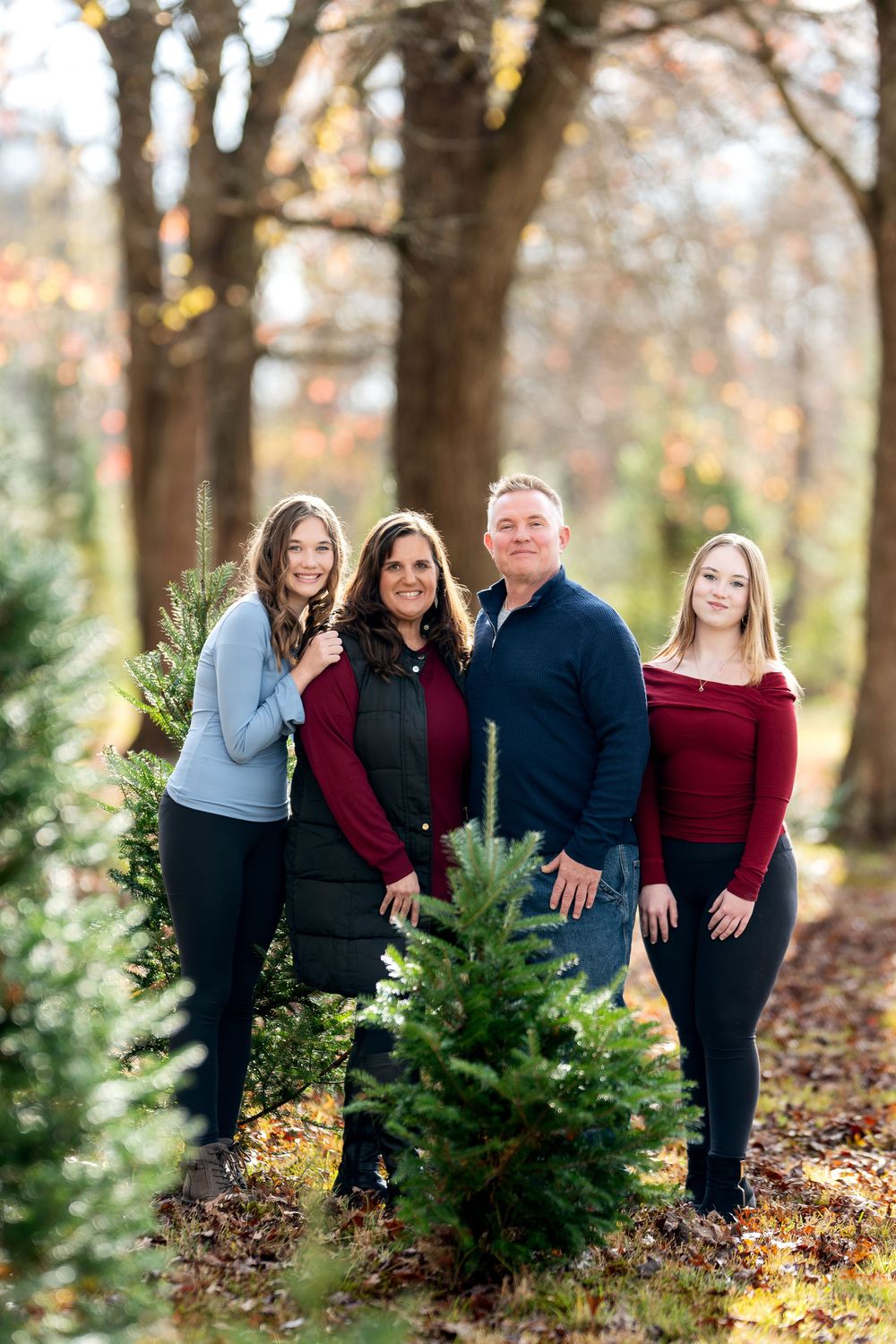Family of four posing together beside Christmas trees in an autumn tree farm with warm fall colors.