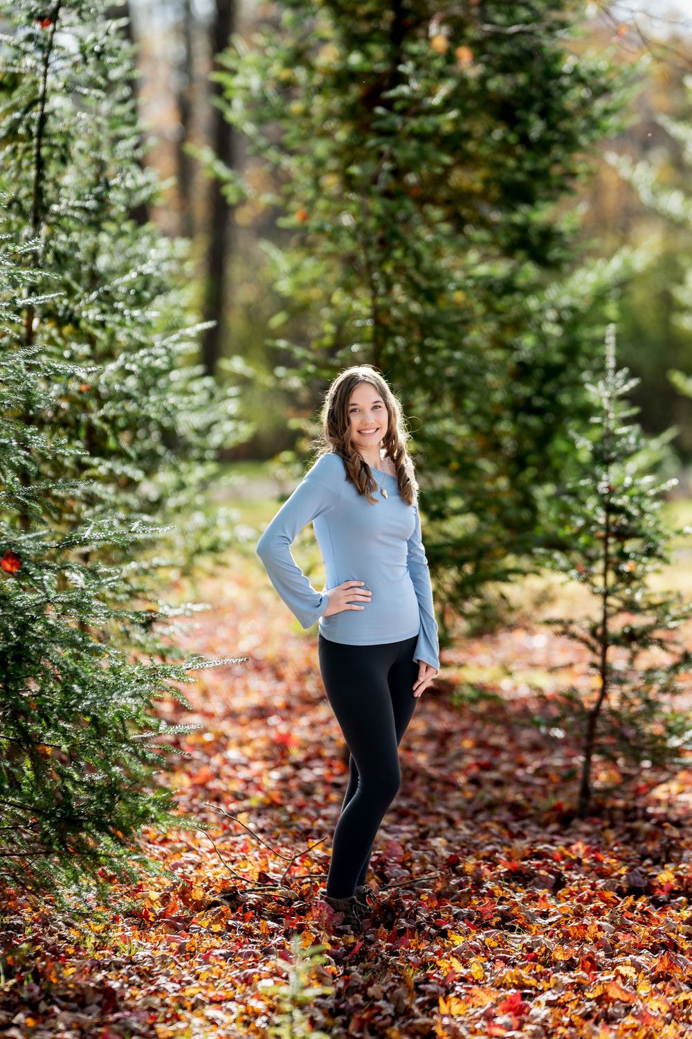Young woman in light blue top posing confidently among pine trees on colorful fall leaves.