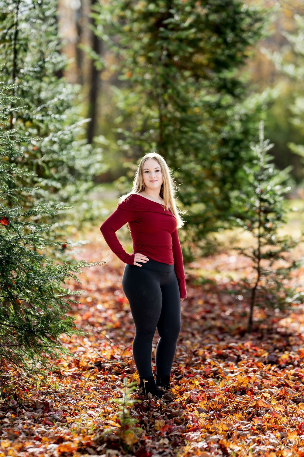 Young blonde woman in red top and black leggings poses among evergreen trees on autumn leaves.