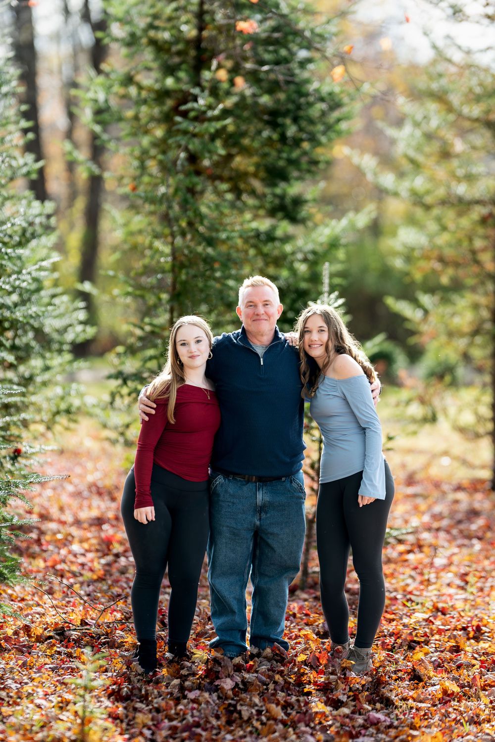 Father posing proudly with his two daughters among pine trees on a carpet of fall leaves.