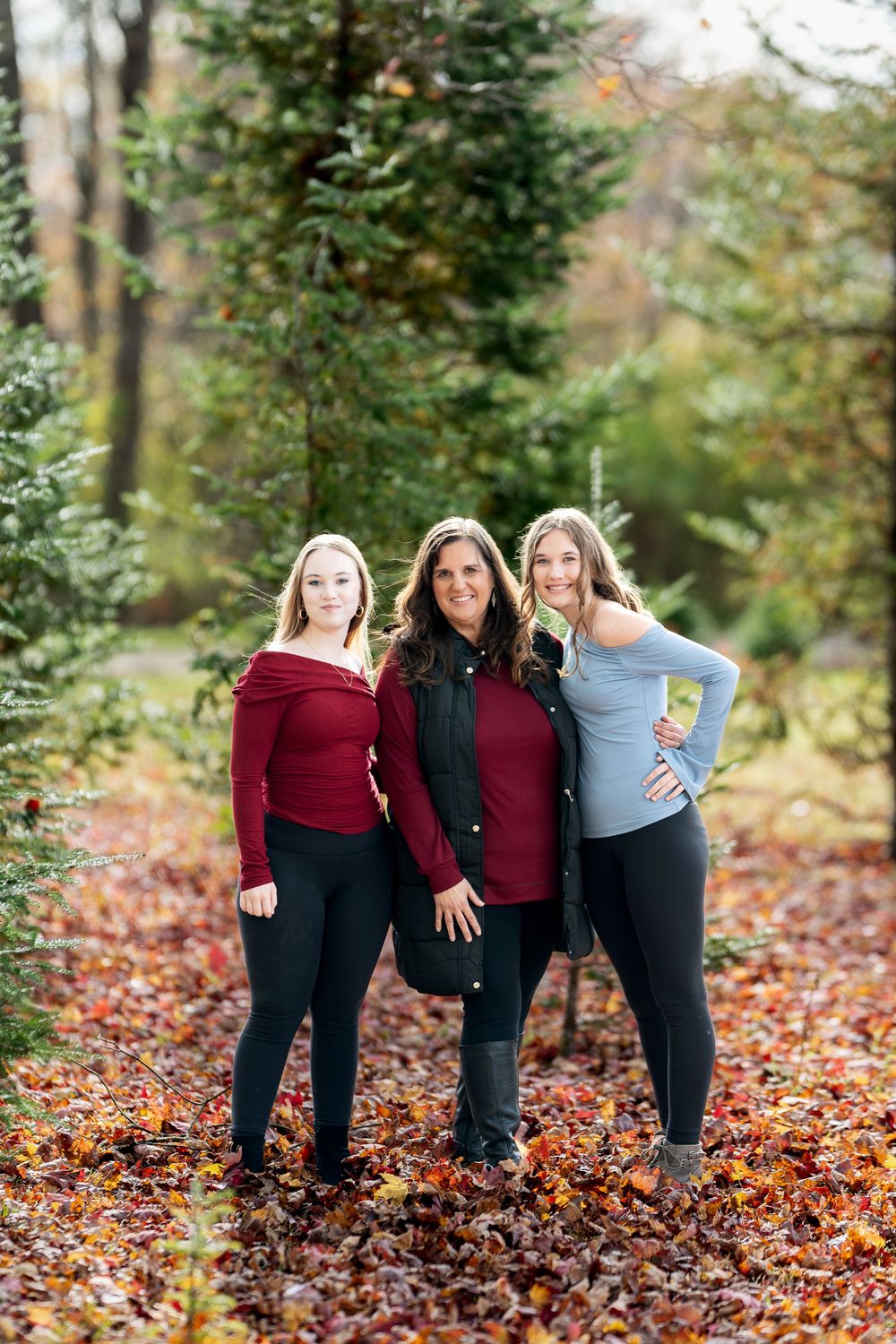 Mother and two daughters posing together among evergreen trees on vibrant autumn fallen leaves.